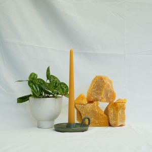 chunks of beeswax next to a beeswax taper candle in a handmade pottery candle holder and a plant